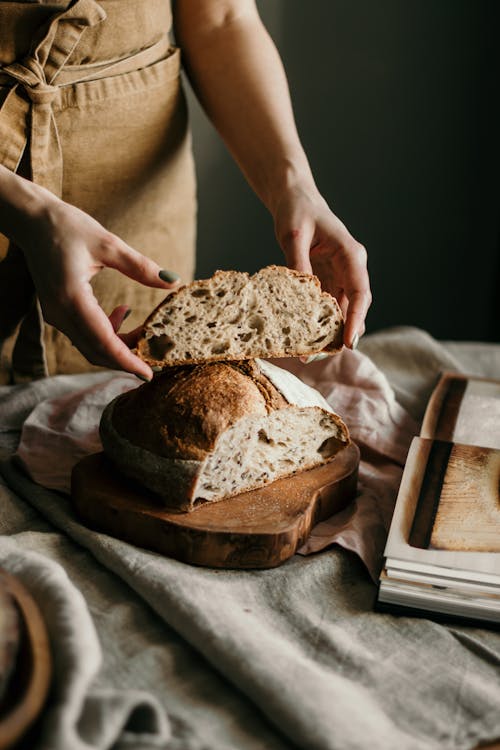 Free High angle of crop anonymous female baker in apron with piece of bread made with recipe from book at home Stock Photo