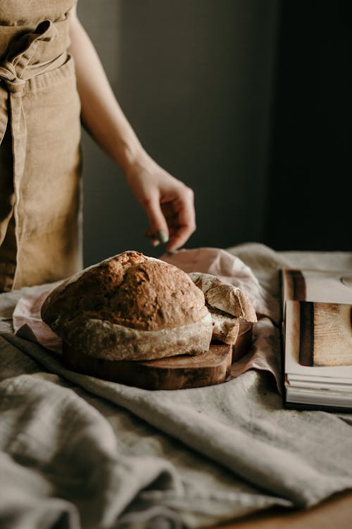 Free Crop anonymous female cook in apron standing at table with freshly baked sourdough bread on cutting board placed on cloth near opened recipe book Stock Photo