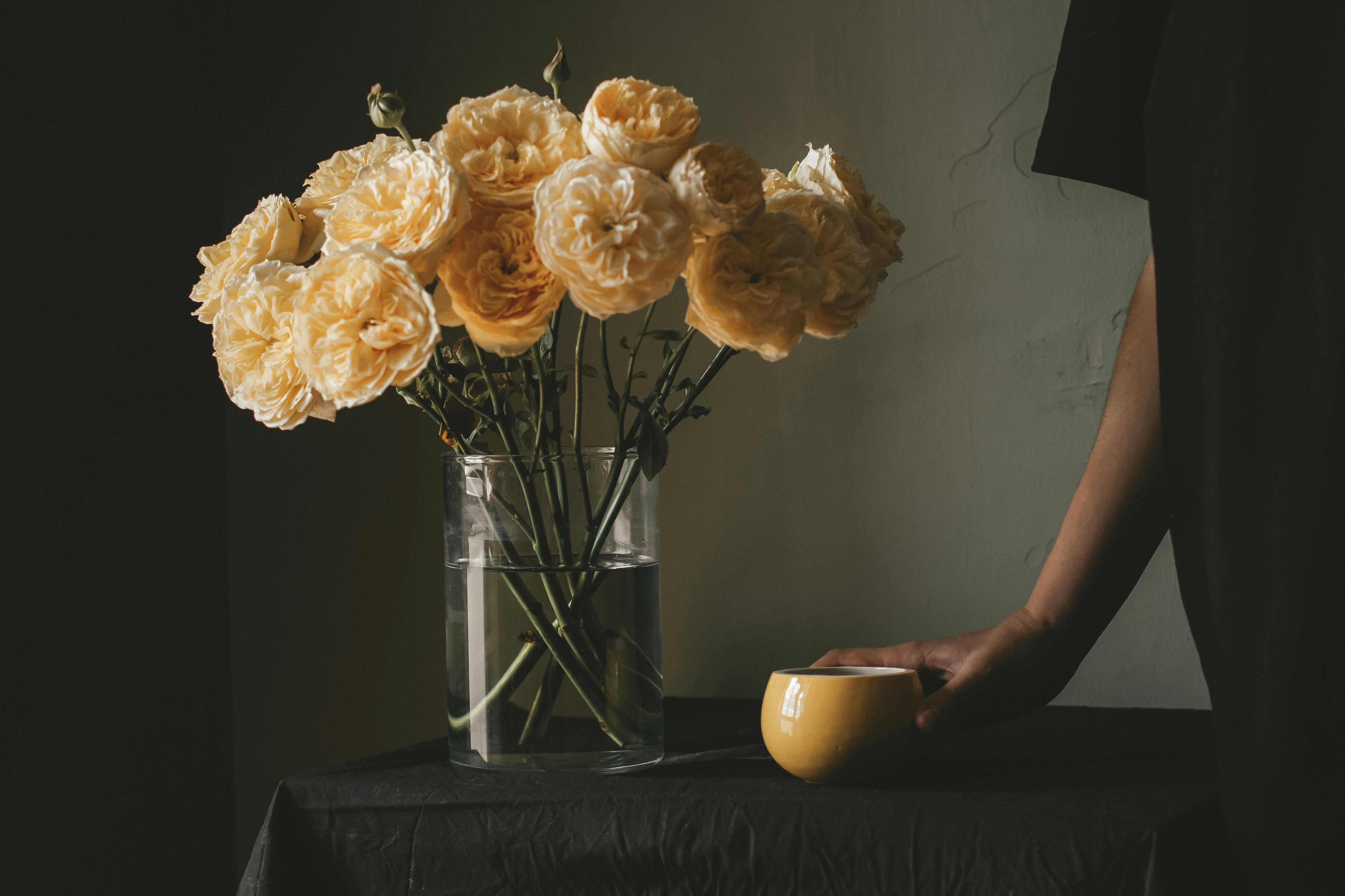 Unrecognizable person standing at table with cup of fresh coffee and glass vase with blooming roses in room at home