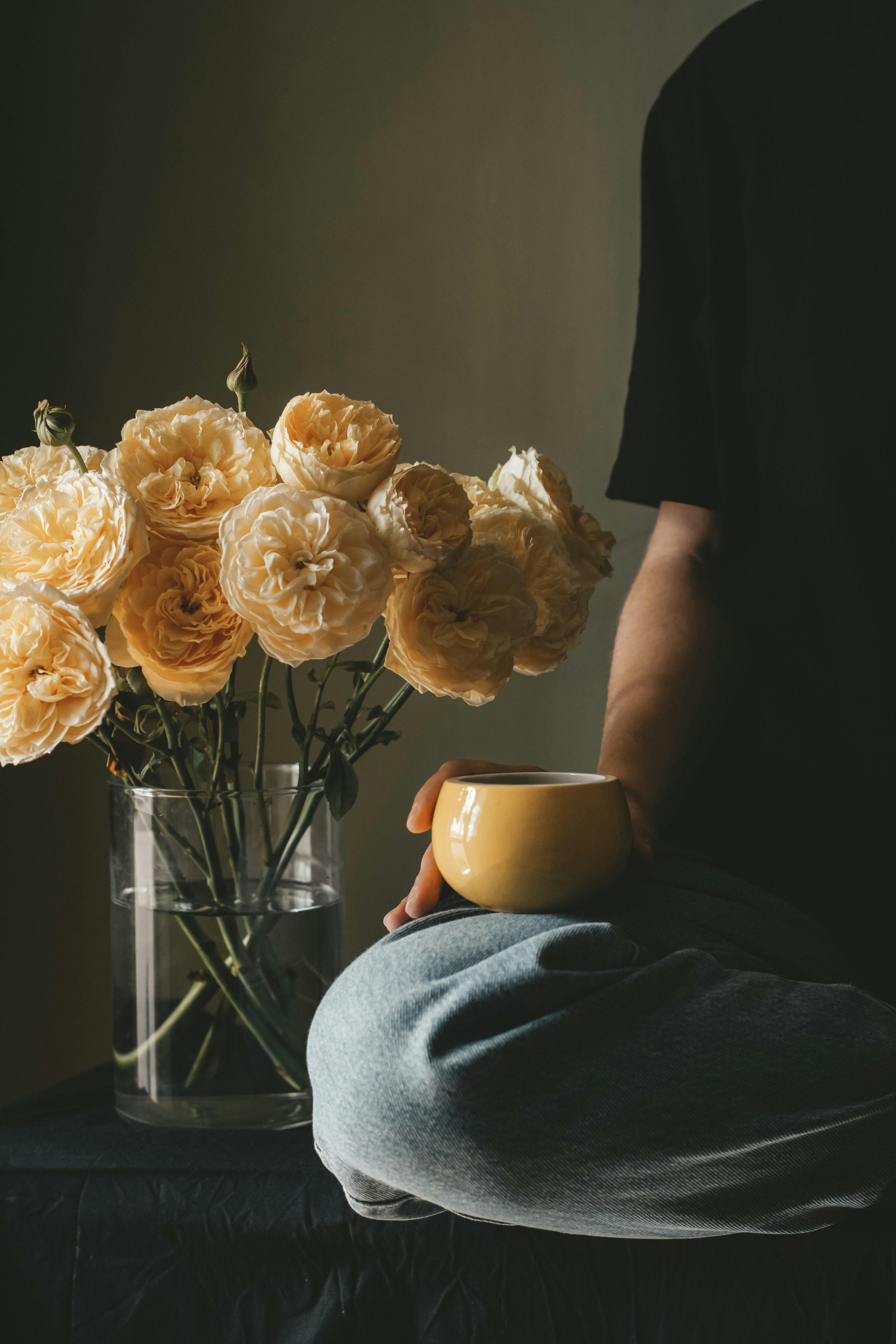Unrecognizable person with cup of hot coffee sitting with crossed legs near vase with blooming roses near wall in room