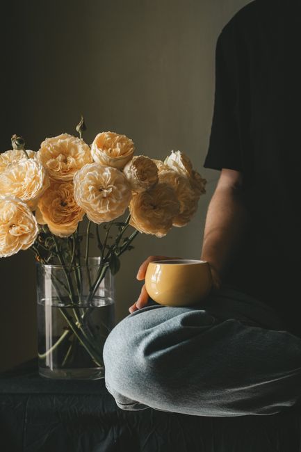 Unrecognizable person with cup of hot coffee sitting with crossed legs near vase with blooming roses near wall in room