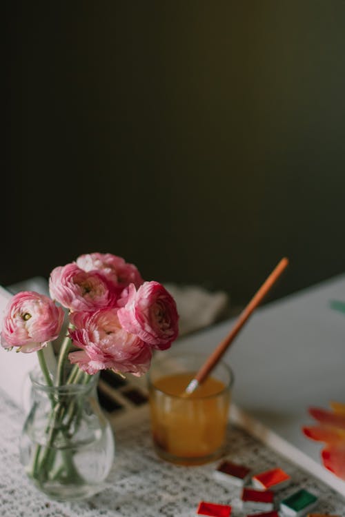 Collection of colorful paints near glass with paintbrush placed on table with painting and vase with blooming flowers in art studio