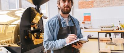 A small business owner in an apron takes notes next to a large coffee roasting machine in a bright workshop.