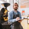A small business owner in an apron takes notes next to a large coffee roasting machine in a bright workshop.