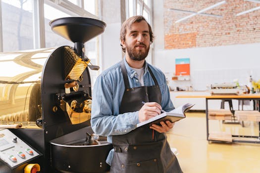 Bearded man in coffee shop taking notes, wearing apron next to roaster, smiling.