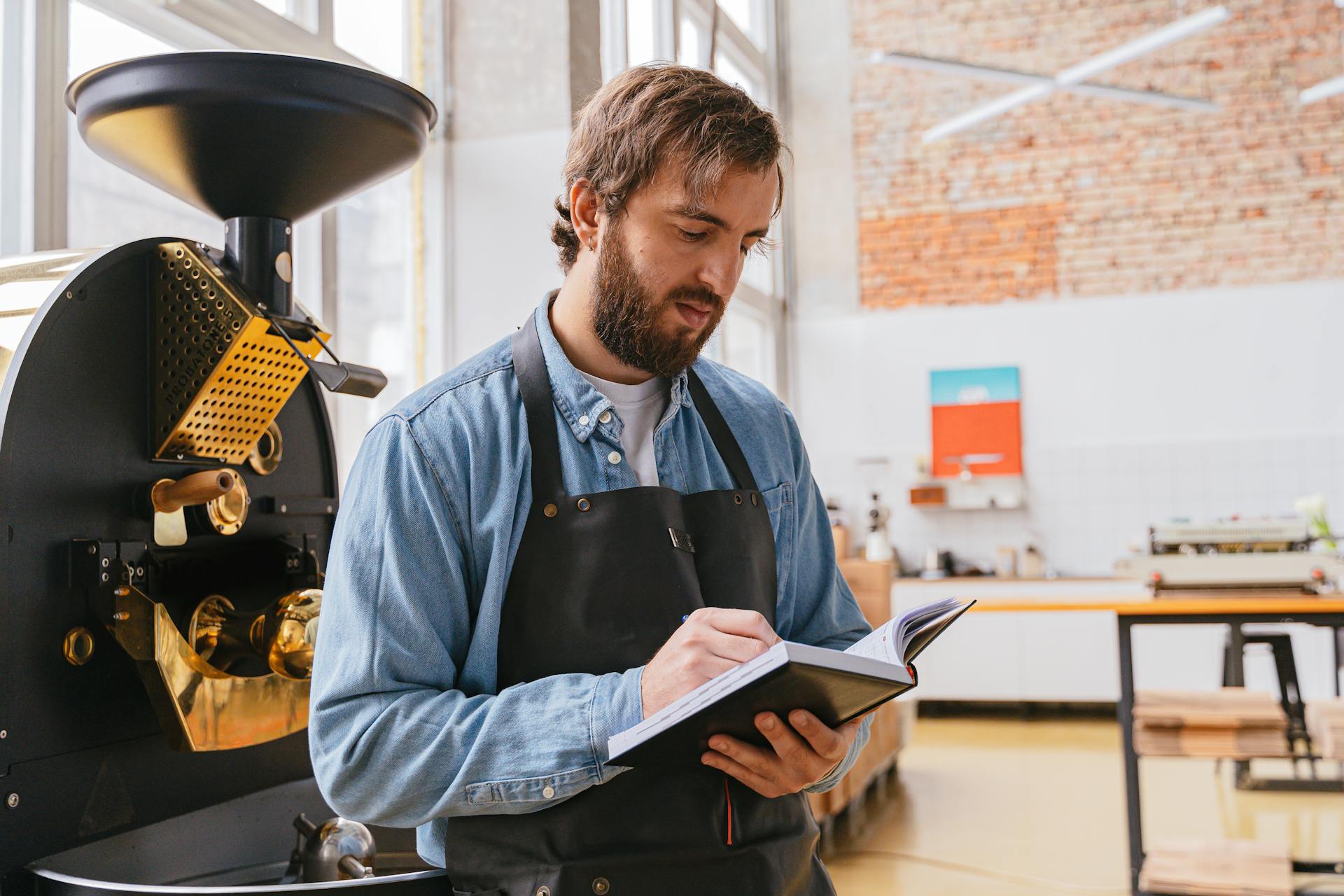 A bearded man in an apron notes observations next to a coffee roasting machine in a bright cafe.