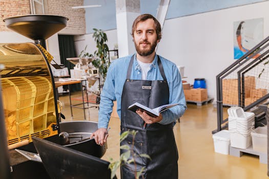 Bearded barista holding notebook while standing next to coffee roaster in contemporary cafe.