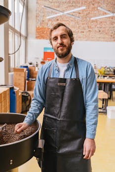 Bearded barista in apron standing by coffee roaster in industrial-style coffee shop.