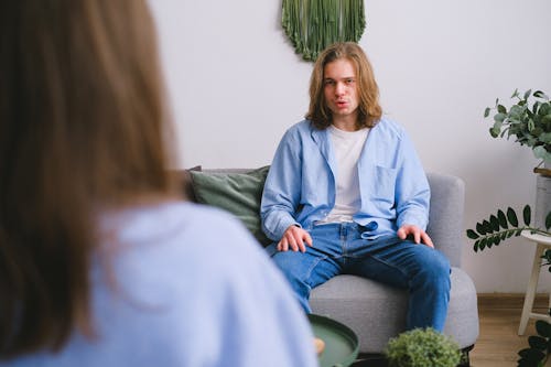Concerned male client siting on sofa while having conversation about problems with faceless psychologist during psychotherapy appointment in light office