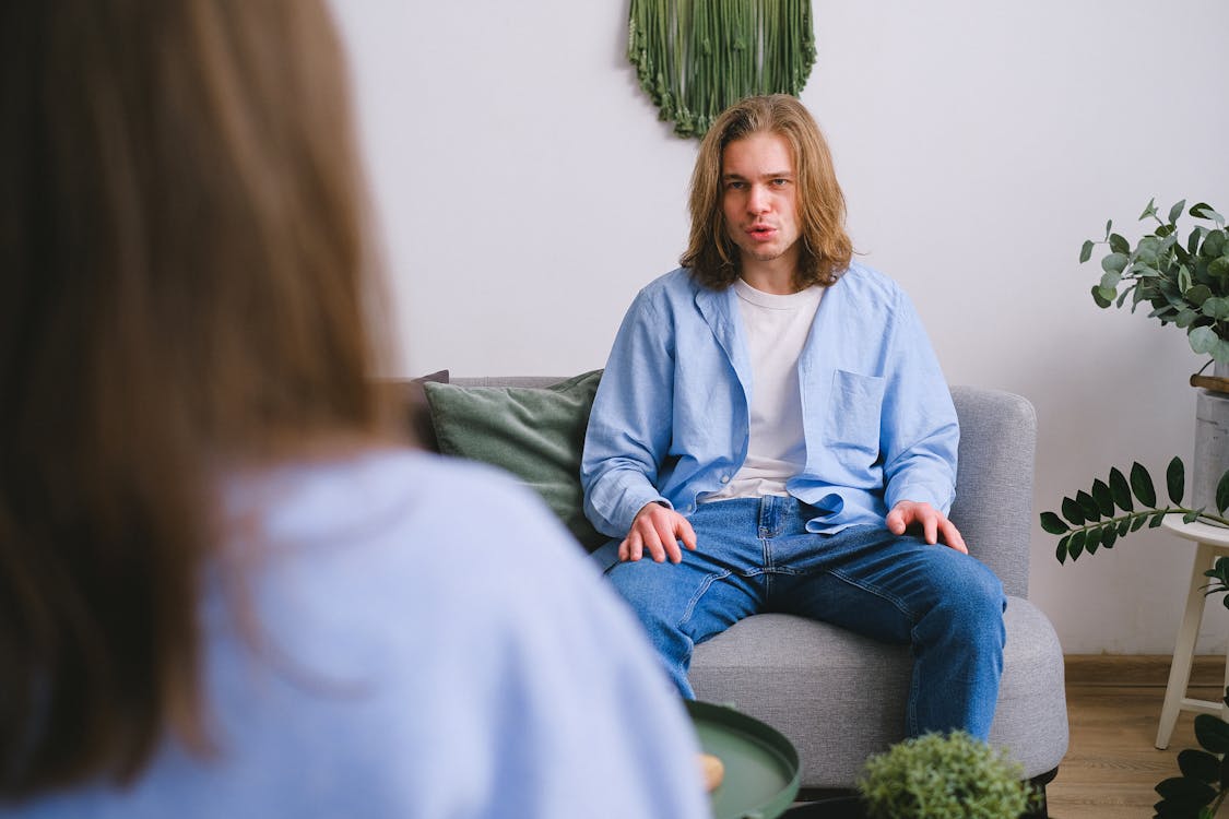 Free Concerned male client siting on sofa while having conversation about problems with faceless psychologist during psychotherapy appointment in light office Stock Photo