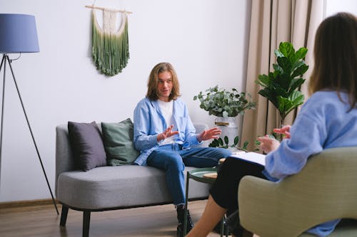 Free Worried male patient sitting on couch while having conversation about problems with unrecognizable professional psychologist during psychotherapy appointment in office Stock Photo