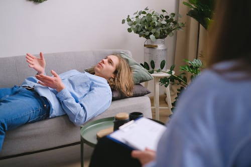 Concerned male client lying on sofa and speaking about mental problems with unrecognizable psychologist with clipboard during psychotherapy appointment in office
