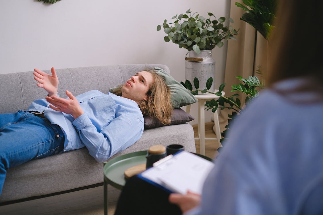 Free Concerned male client lying on sofa and speaking about mental problems with unrecognizable psychologist with clipboard during psychotherapy appointment in office Stock Photo