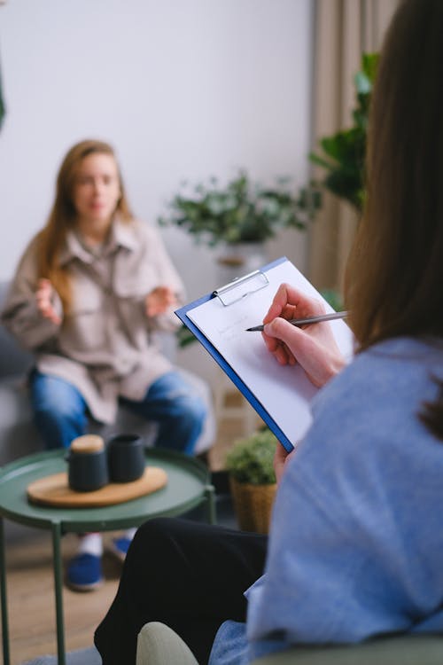 Free Unrecognizable female psychologist taking notes on clipboard while listening to patient sitting on blurred background during psychotherapy consultation in office Stock Photo