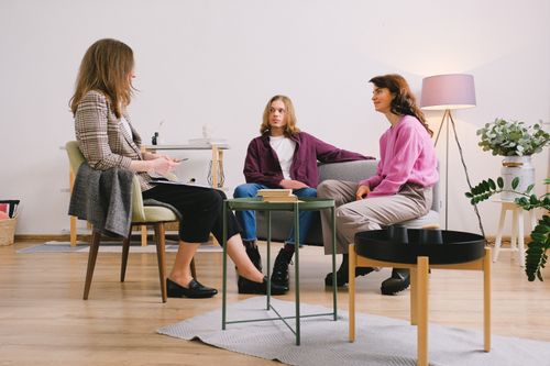 Free Three people engaged in conversation in a cozy, modern living room setting with warm lighting. Stock Photo