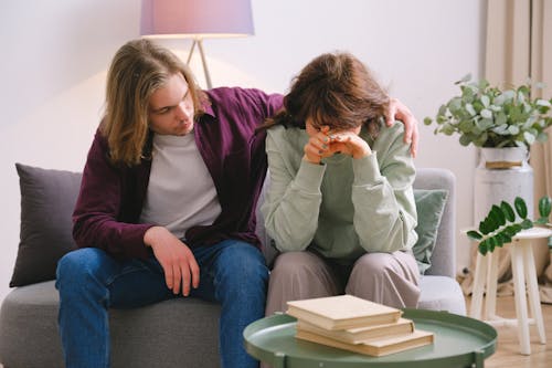 Free Young male in jeans comforting depressed upset female crying on gray sofa near table with books Stock Photo