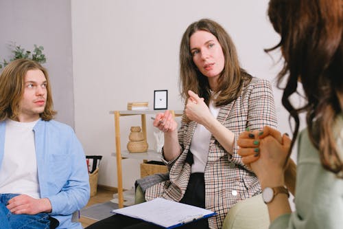 Thoughtful woman psychologist in stylish clothes siting with clipboard and talking with couple in light room