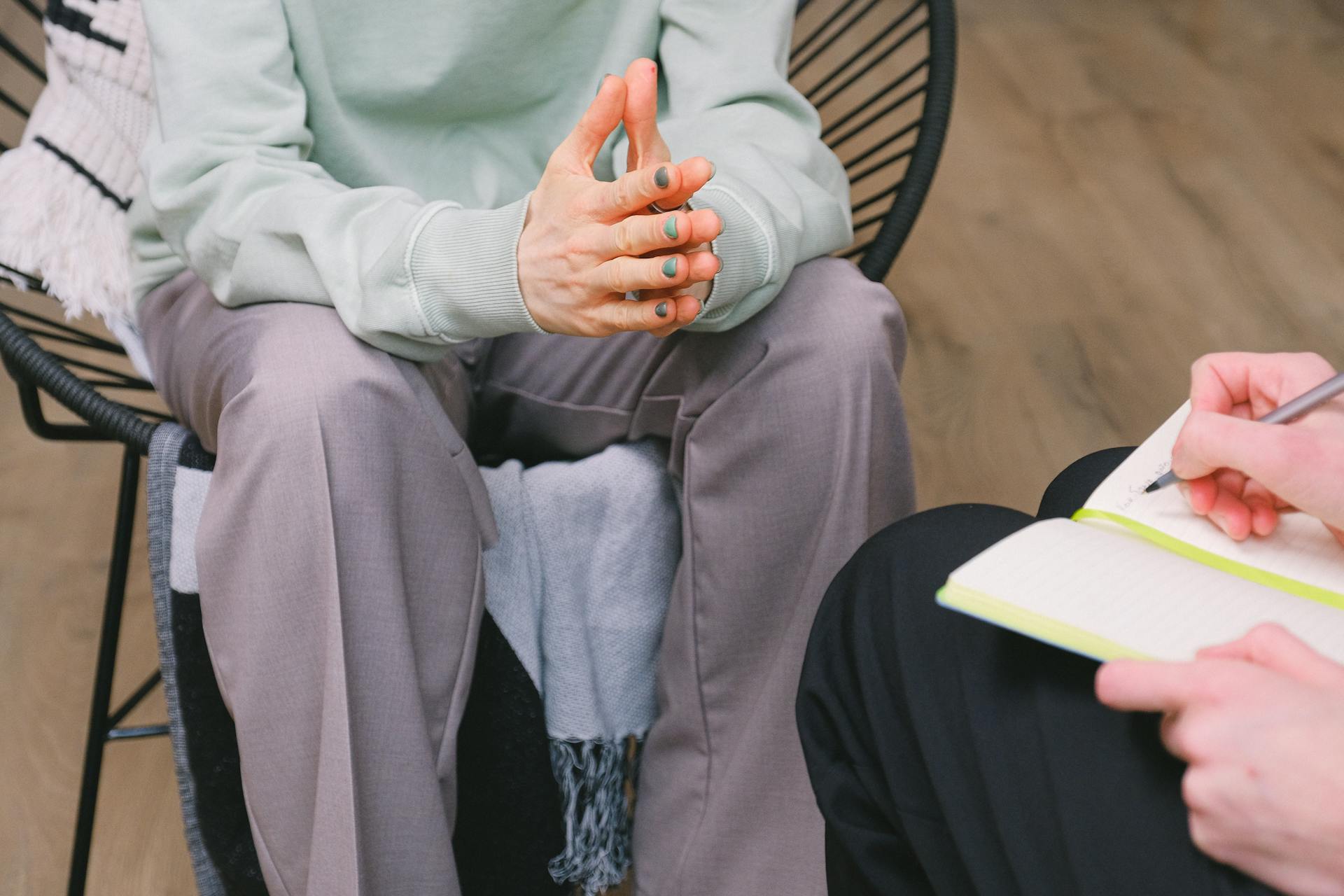 Crop unrecognizable psychotherapist taking notes in journal while interacting with woman on chair during psychological counseling indoors