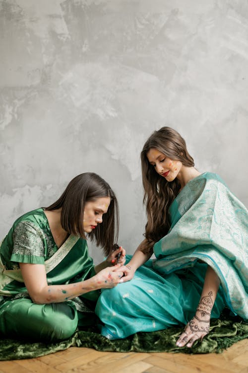 Woman in Green Sari Dress Sitting Beside Woman in Teal Sari Dress and Painting Mehendi Pattern on Hand