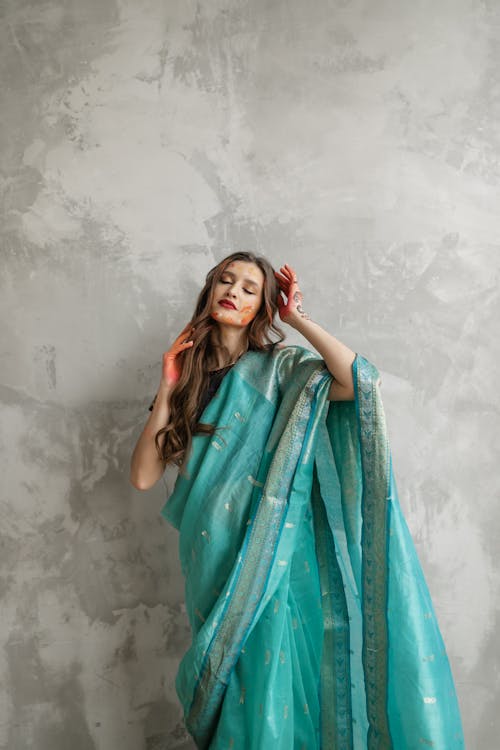 Woman in Teal Sari Dress With Eyes Closed Standing Behind Gray Wall with Hands Near Face