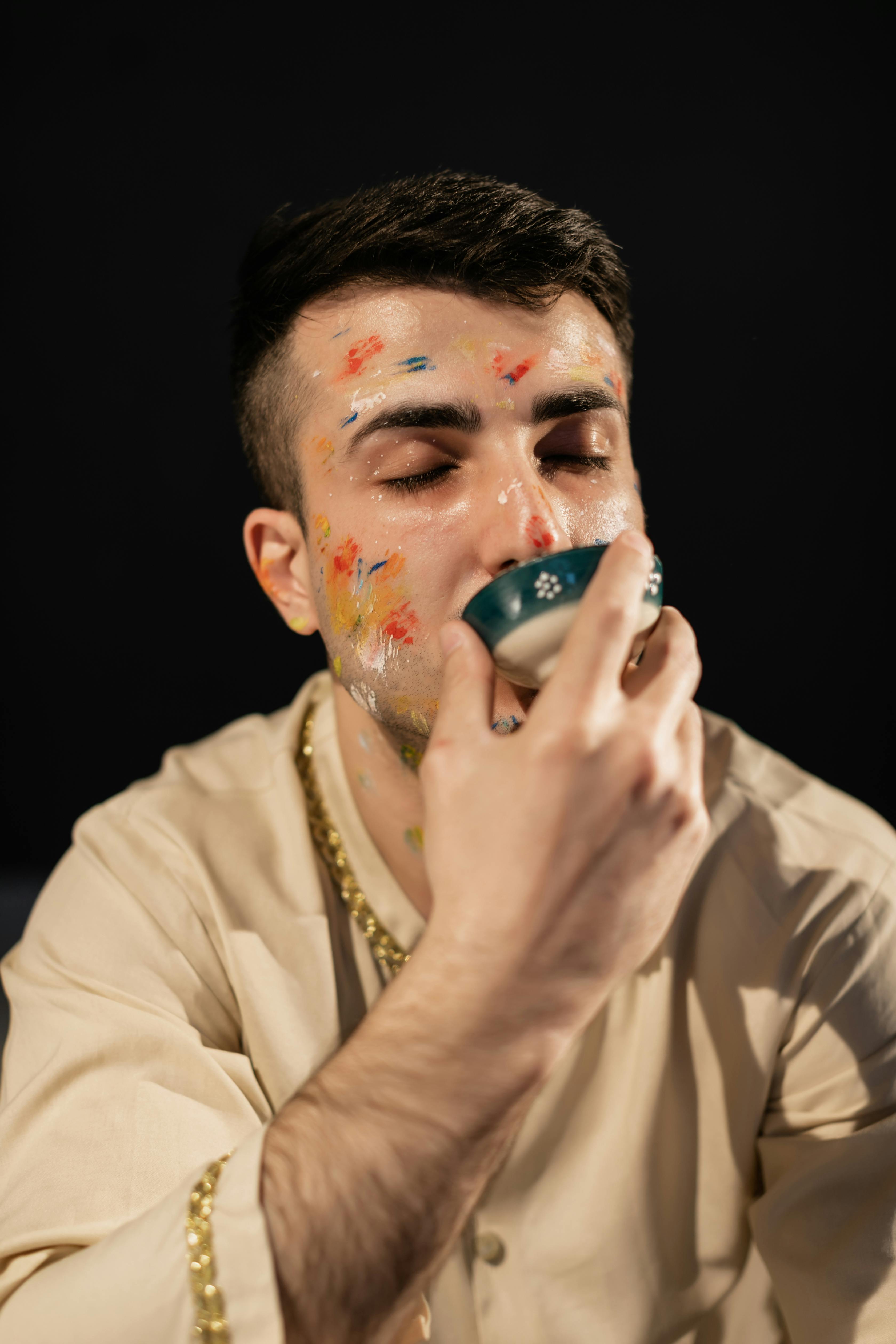 A man with colorful face paint is drinking from a cup, celebrating Holi.