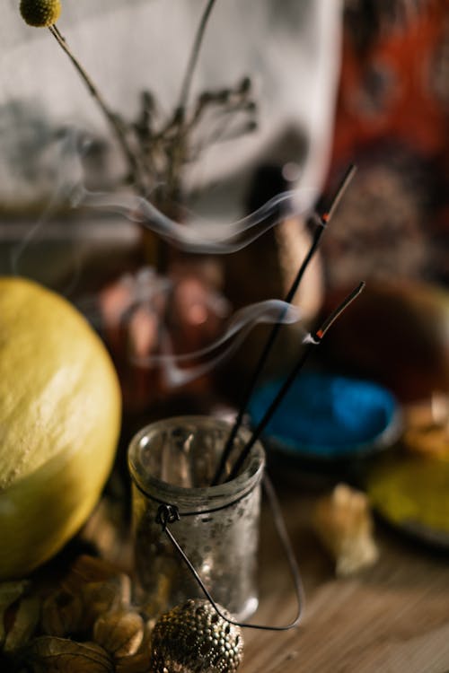Burning incense sticks with wavy smoke spreading in air above table with assorted decor on blurred background
