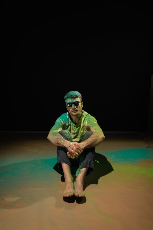 Free A man sitting on the floor, covered with vibrant Holi powder against a dark background. Stock Photo