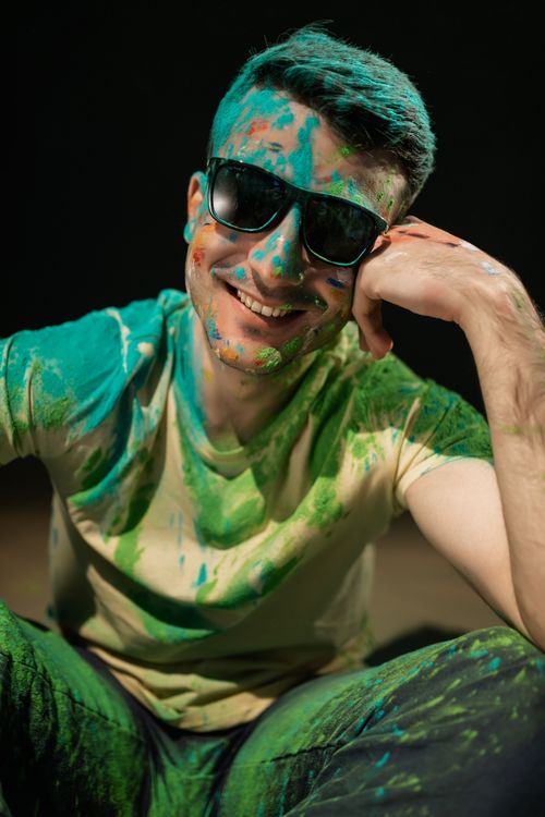 Free A cheerful man wearing sunglasses, covered in vibrant Holi powder, sits smiling. Stock Photo