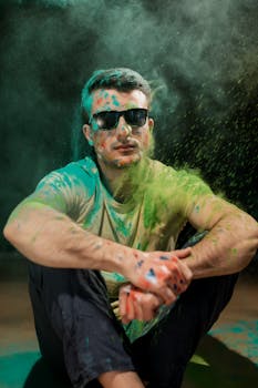 A young man wearing sunglasses covered in colorful Holi powder, sitting and posing.