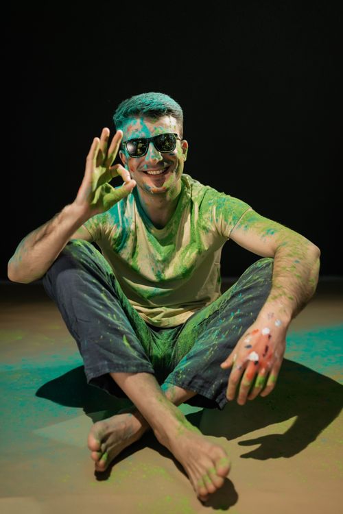 Free A smiling man covered in vibrant Holi powder sitting indoors making an okay hand gesture. Stock Photo