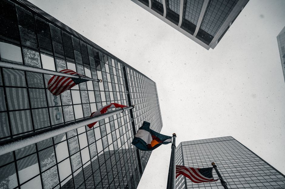 Looking up at flags and tall glass buildings during snowfall in downtown Denver, CO.