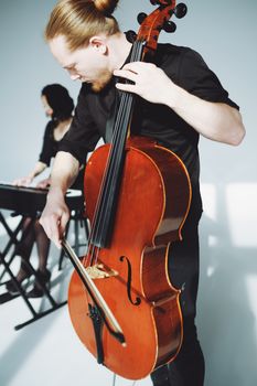 A male musician with a man bun skillfully playing a cello, accompanied by a keyboard player.