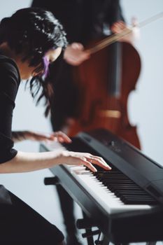 A close-up of a woman playing the keyboard with a blurred background featuring a cello.