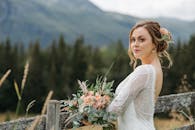 一位美丽的新娘拿着花束在质朴的木栅栏和风景秀丽的山脉背景下摆姿势