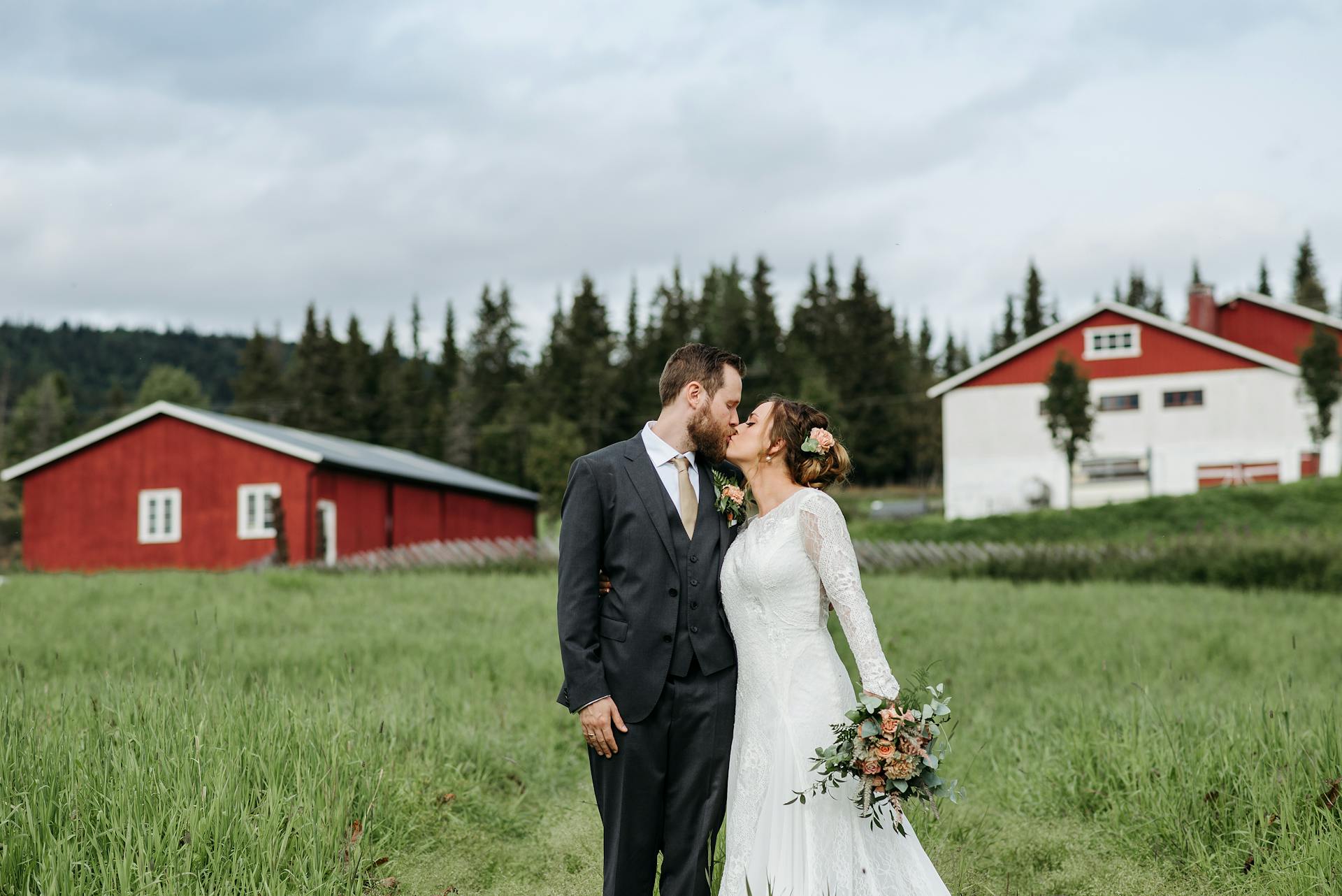 Newlyweds share a kiss in a scenic field with rustic barns and trees.