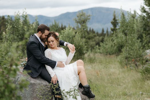 A couple in wedding attire sits together on a rock amidst lush greenery, capturing a serene and romantic moment.