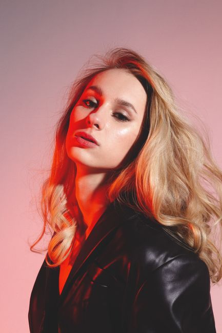 Confident young woman in leather black jacket looking at camera in bright studio with red lights