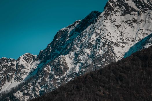 Steep slope of rocky mountains with uneven surface covered with hoarfrost located near dense woods with coniferous trees on cold winter day