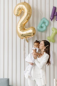 A loving mother holds her child under a golden number two balloon for a birthday celebration indoors.