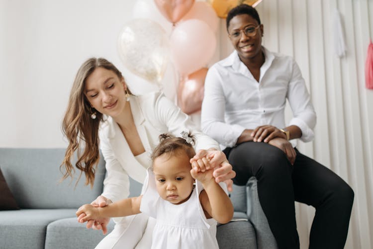 Woman In White Blazer Holding Hands Of Baby Girl In White Dress Walking While Man In White Shirt Sitting On Sofa