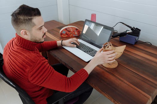 Engineer working on a laptop at a wooden desk, holding a 3D model in an office setting.