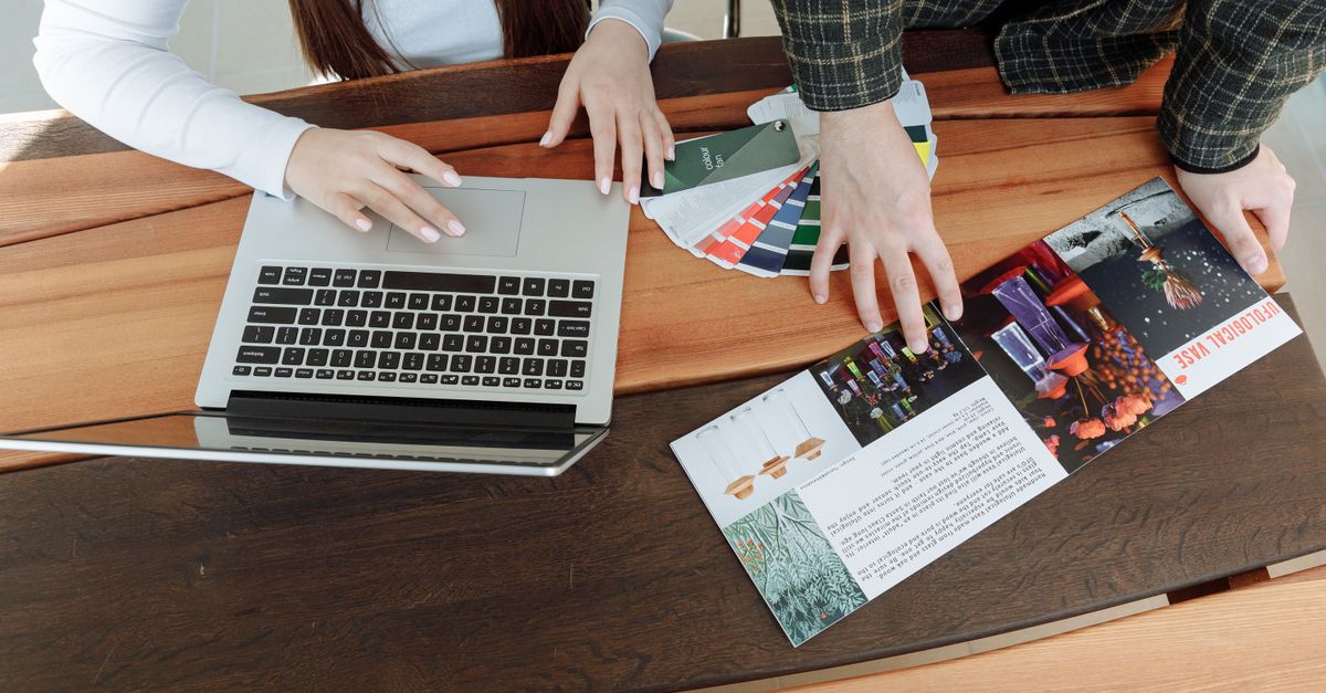 Two colleagues working on a design project using a laptop and color guides at a wooden table.