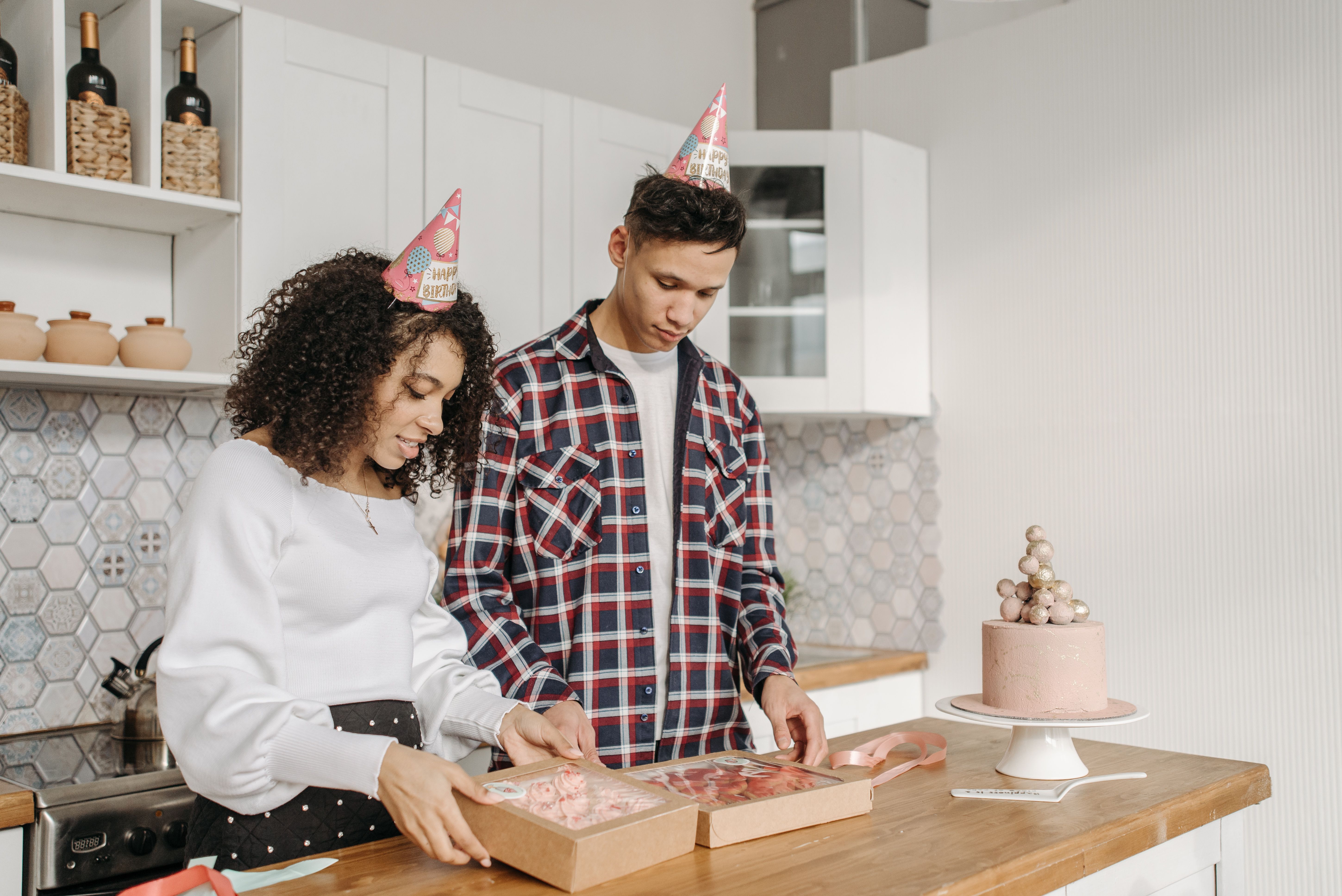 Jovens comemorando aniversário em um ambiente de cozinha, com chapéus de festa e presente, celebrando uma ocasião especial em casa.