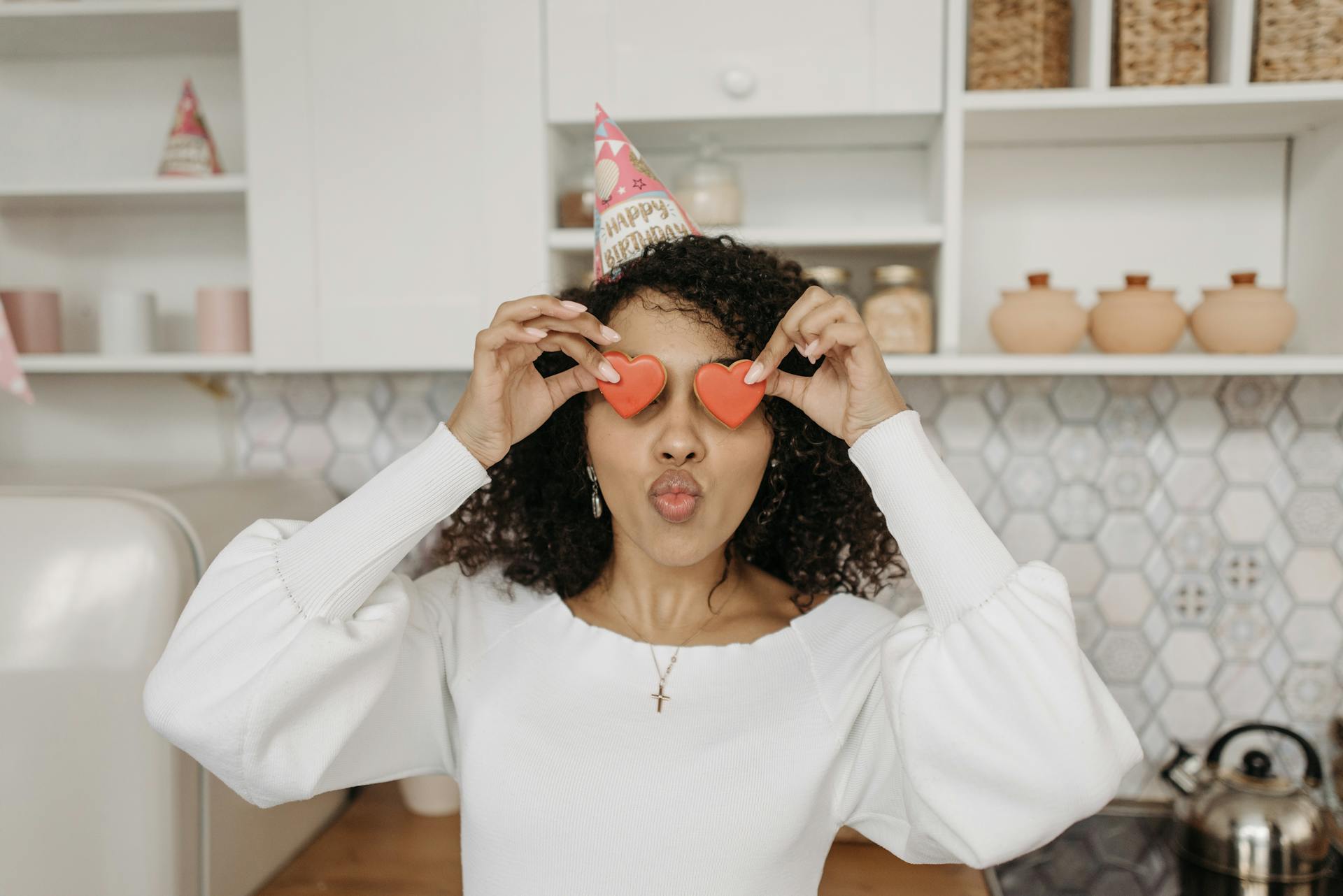 Young woman celebrating birthday with heart-shaped props and party hat in a bright kitchen.