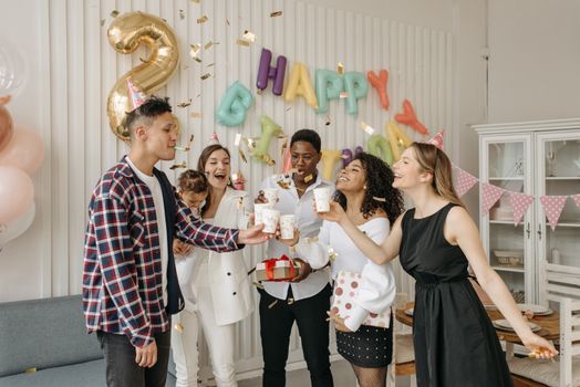 A diverse group of friends celebrating a birthday indoors with balloons and party hats.