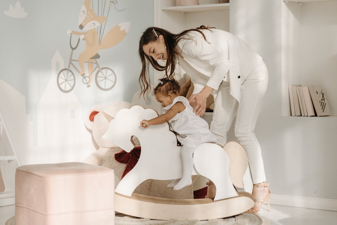 Free Woman in White Clothes Helping a Child Ride a Rocking Horse Stock Photo