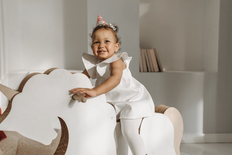 Baby Girl In White Dress Sitting On Rocking Horse