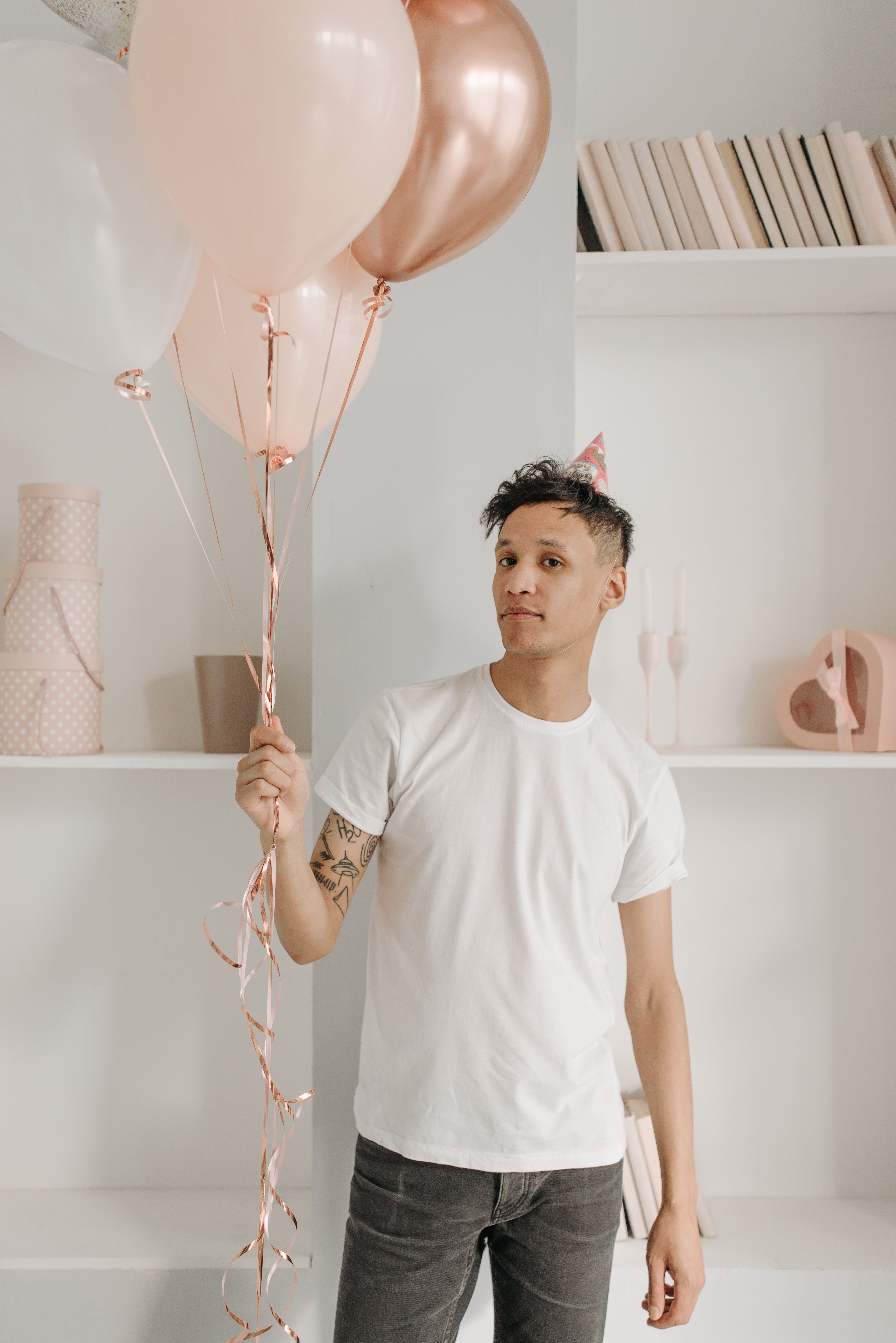 Young man in a white shirt holding balloons at an indoor birthday party, wearing a party hat.