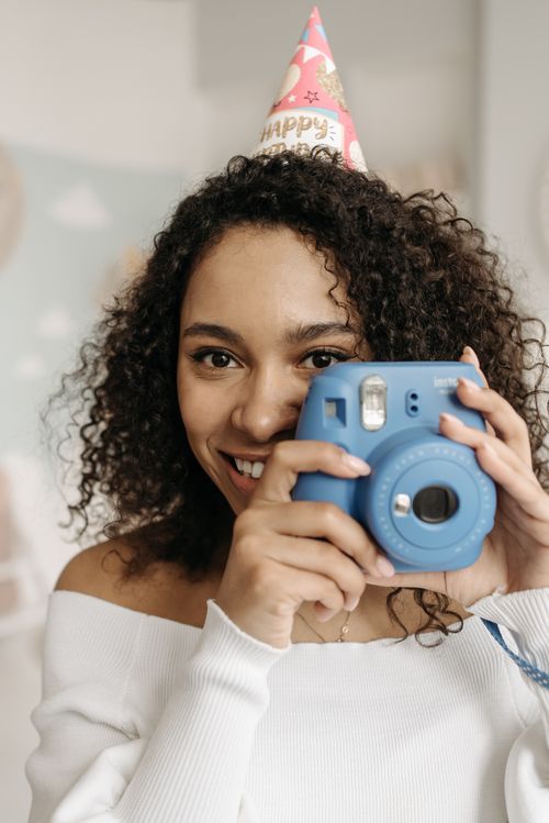 Free A cheerful woman with curly hair and a party hat holding a blue polaroid camera indoors. Stock Photo