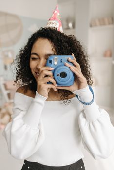Smiling woman with curly hair in party hat taking photo indoors with a vintage camera.